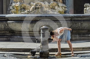 Young charming girl tourist drinks water from an antique drinking fountain of the roman nose against the background of historical