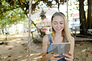 Young charming girl using tablet and riding swing on sand, wearing jeans sundress.