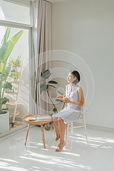 Young charming woman sitting on chair and reading book next to window at home