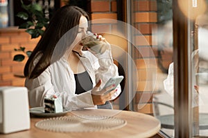 Young charming woman is using cell phone while having coffee break while sitting in cafe in free time, attractive woman