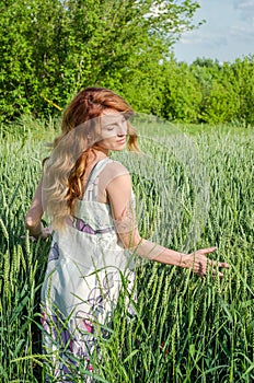 Young charming woman walking outdoors in a field near the green bushes and trees, hand patting wheat ears, dressed in a beautiful