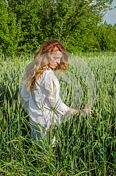 Young charming woman walking outdoors in a field near the green bushes and trees, hand patting wheat ears, dressed in a beautiful