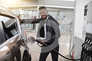 Young cheerful African American businessman refueling his modern luxury car at fuel station, smiling to camera