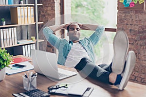 Young cheerful afro freelancer is resting at a workplace, with feet on top of the desk, with closed eyes, smiling, dreaming