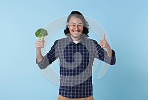 Young cheerful asian bearded man showing broccoli and hand thumbs up isolated on blue background