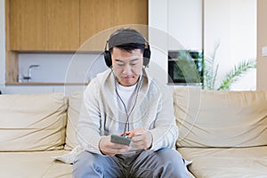 cheerful asian guy listening to music in headphones sitting on sofa at home. A man alone on the couch feels good lifestyle.