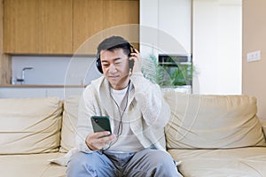 cheerful asian guy listening to music in headphones sitting on sofa at home. A man alone on the couch feels good lifestyle.