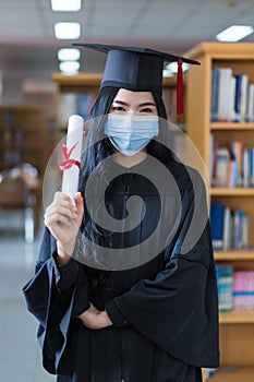 A young cheerful Asian woman university graduate in graduation gown and cap wears a sanitary mask holds a degree certificate to