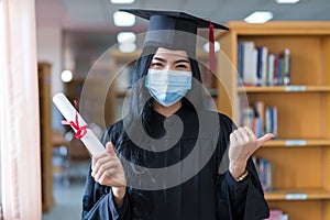 A young cheerful Asian woman university graduate in graduation gown and cap wears a sanitary mask holds a degree certificate to