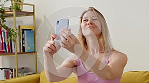 young cheerful blonde woman in pink top sits on couch at home and takes selfie on her blue phone. Happy woman is