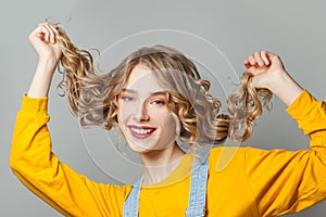 Young cheerful blonde woman touching her long curly hair on gray background