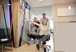 Young cheerful business people in smart casual wear having fun while racing on office chairs and smiling.