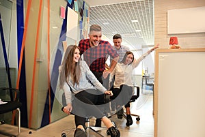 Young cheerful business people in smart casual wear having fun while racing on office chairs and smiling.