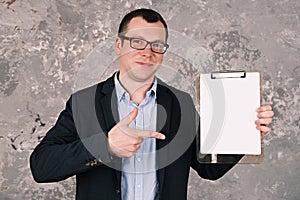 Young cheerful businessman or manager in glasses and suit holding clipboard with blank sheet of paper in his hands and
