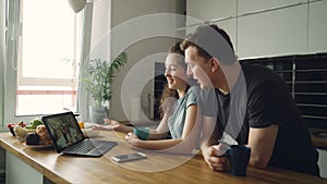 Young cheerful caucasian couple sitting at table in front of laptop skyping with their happy positive tanned friends