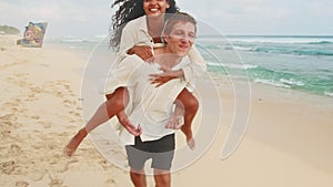 Young cheerful Caucasian man rides Indian woman on back located on sea beach