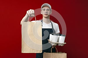 Young cheerful courier in uniform gives a parcel and holds an order, delivery man holds a package on a red background