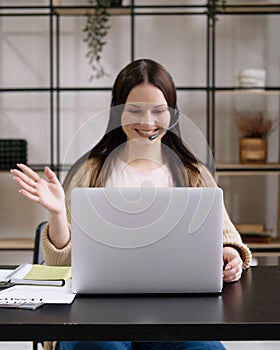 Young cheerful cute beautiful business woman sit indoors in office using laptop computer with headset.