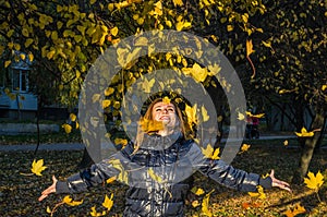 Young cheerful cute girl woman playing with fallen autumn yellow leaves in the park near the tree, laughing and smiling