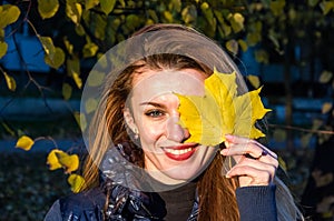 Young cheerful cute girl woman playing with fallen autumn yellow leaves in the park near the tree, laughing and smiling