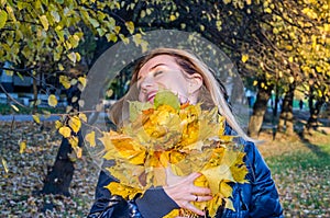 Young cheerful cute girl woman playing with fallen autumn yellow leaves in the park near the tree, laughing and smiling