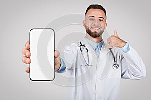 Young cheerful doctor showing phone with blank screen and gesturing call me