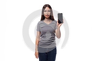 young cheerful european brunette woman dressed in gray basic t-shirt holding smartphone with screen mockup