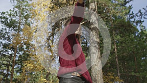 Young cheerful female dressed in red knitted jumper gives thumbs up looking at the sun in autumn forest. Low angle 4K