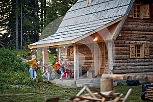 Young cheerful friends in front of wooden cottage on the terrace.  Guy and girl playfully fighting over beer. Girl playing guitar