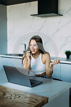 Young cheerful happy woman indoors at home at the kitchen using laptop computer talking by mobile phone