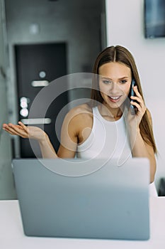 Young cheerful happy woman indoors at home at the kitchen using laptop computer talking by mobile phone
