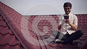 Young cheerful hipster sitting on house roof with smartphone in hands, playing video games, wearing round sunglasses and shirt.