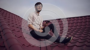 Young cheerful hipster sitting on house roof with smartphone in hands, wearing round sunglasses and shirt.