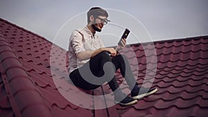 Young cheerful hipster sitting on house roof with smartphone in hands, wearing round sunglasses and shirt.
