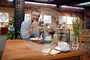 Young cheerful man at cafe smiling and looking at phone. Enjoying cup of coffee and cake