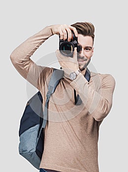Young cheerful man photographer takes images with digital camera. Isolated on gray background