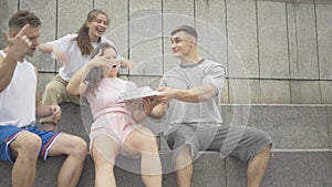 Young cheerful man with pizza jumping up to group of friends sitting on urban stairs. Portrait of joyful Caucasian