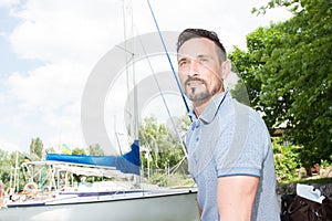 Young cheerful man relaxing on sailboat posing and looking at far on background of boats, sky and trees.