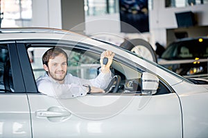 Young cheerful man showing key in his car in dealership