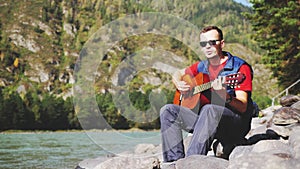 Young cheerful man wearing sunglasses plays on a guitar sings song sits next to mountain river on sunny day.