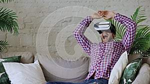 Young cheerful man wearing virtual reality headset having 360 VR video experience while sitting on couch in living room