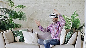 Young cheerful man wearing virtual reality headset having 360 VR video experience while sitting on couch in living room
