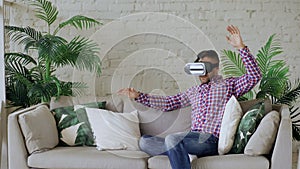 Young cheerful man wearing virtual reality headset having 360 VR video experience while sitting on couch in living room