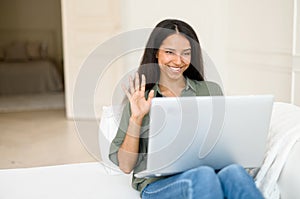 Young cheerful multiethnic woman waving hello to online interlocutor, using laptop