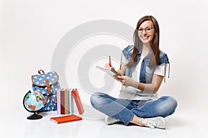 Young cheerful smiling woman student in glasses writing notes on notebook sitting near globe, backpack, school books