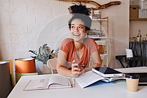 Young cheerful student girl with dark curly hair sitting at the table with laptop and cup of coffee to go holding pencil