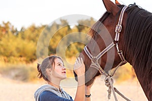 Young cheerful teenage girl stroking brown horse`s nose. Outdoor
