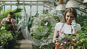 Young cheerful woman in apron and gloves talking phone while gardening plants and loosen ground in flower in greenhouse