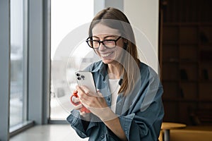 A young cheerful woman in glasses looks at the smartphone screen and laughs.