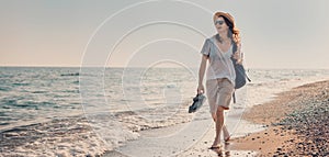 Young cheerful woman in a hat and sunglasses walking on the seashore with a backpack at sunset. Summer sea holidays